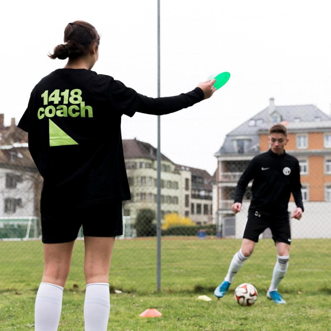 Eine Frau mit 1418-Coach-Trikot steht auf dem Fussballplatz und hält ein Trainings-Hütchen in der Hand. Im Hintergrund trainiert ein Fussballer. 