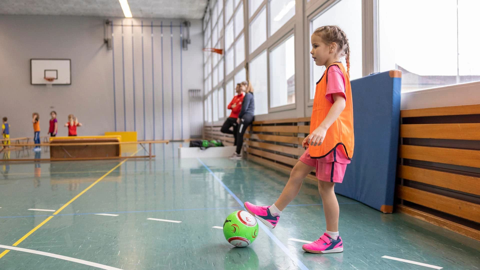 Ein Mädchen mit orangem Trikot steht in einer Turnhalle und hat den Fuss am Ball. 