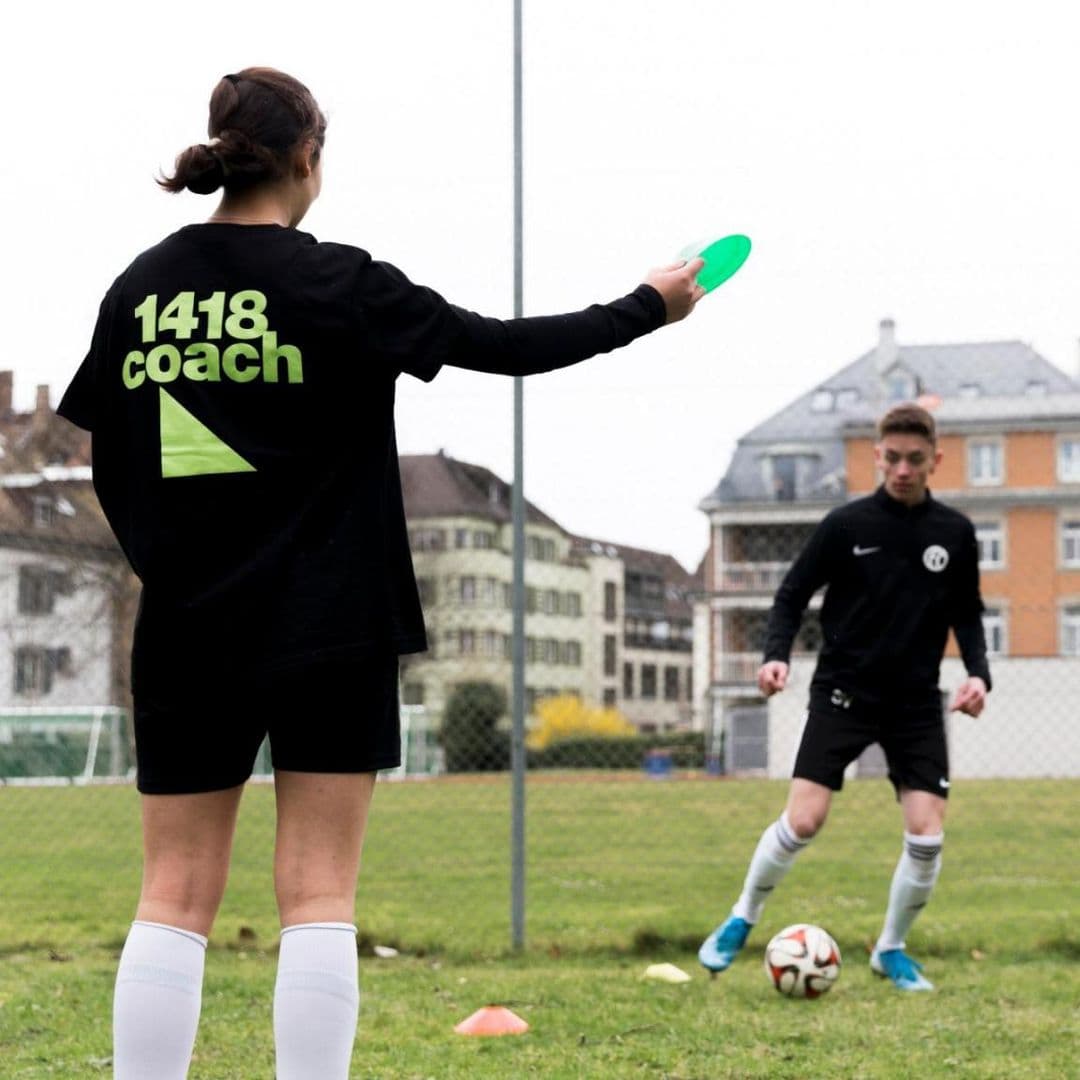 Eine Frau mit 1418-Coach-Trikot steht auf dem Fussballplatz und hält ein Trainings-Hütchen in der Hand. Im Hintergrund trainiert ein Fussballer. 